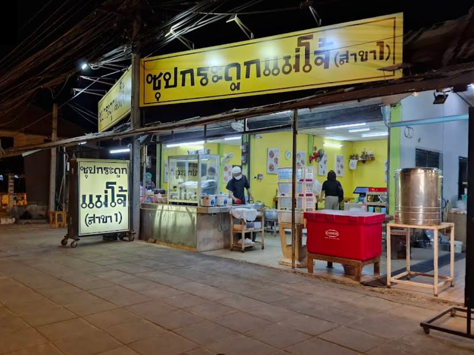 ซุปกระดูกแม่โจ้สาขา1 Nighttime street view of a small Thai noodle shop with bright yellow signage reading ‘ซุปกระดูกแม่โจ้ (สาขา1)’. The shop is open to the sidewalk, with staff preparing food inside under bright lights, a red cooler and large metal pot near the entrance, and overhead power lines visible above the street.